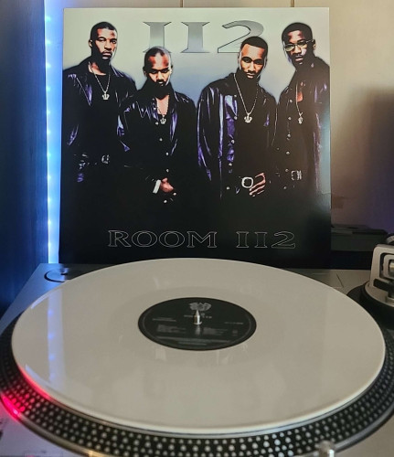 A white vinyl record sits on a turntable. Behind the turntable, a vinyl album outer sleeve is displayed. The front cover shows the 4 members of 112 standing next to each other looking at the camera.