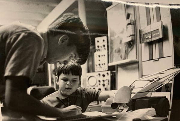 A teenager writes in a notebook hunched over a table as he stands, a younger boy to his left looks on intently. There is computing and oscilloscope, and other electrical equipment and peripherals in the background of this black and white rectangular (landscape orientation photo). The two boys are in focus, the background is not.