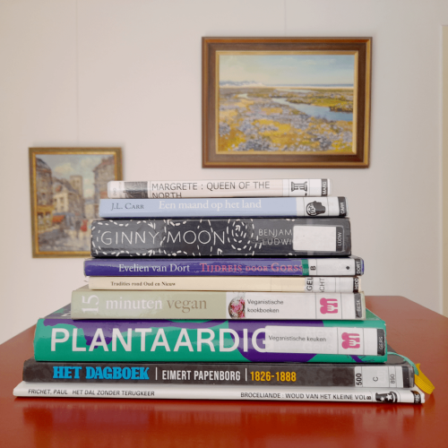 A colourful stack of books on a red surface. In the background are two colourful paintings, one of a street and one of a river running through a field, against a white wall.
