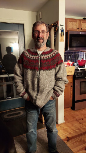 Gray haired bearded white man with a mustache and glasses wearing a handknit Icelandic cardigan. Sweater is gray with the yoke design in red and green.