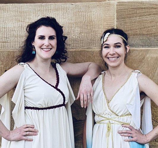 Dr Rad and Dr G smiling while dressed as Roman goddesses in front of a sandstone building.