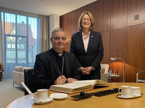 Der Nuntius sitzt an einem Tisch im Büro der Präsidentin und trägt sich ins Gästebuch ein. Auf dem Tisch sind Kaffeetassen eingedeckt. Hinter dem Nuntius steht Präsidentin Antje Grotheer.