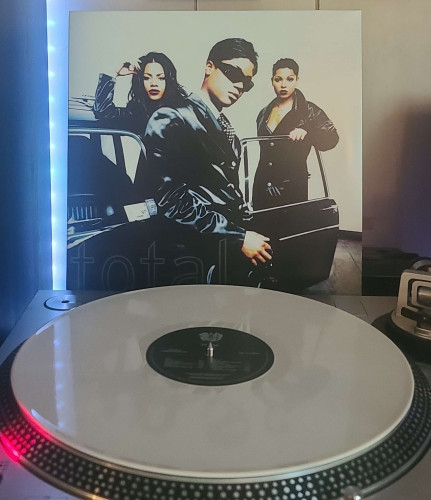A white vinyl record sits on a turntable. Behind the turntable, a vinyl album outer sleeve is displayed. The front cover shows the 3 members of Total standing around a car with its driver's side door open. 