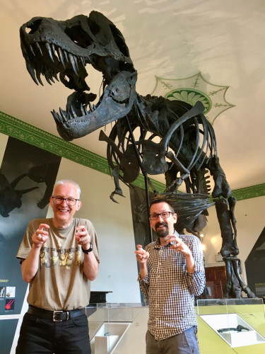Two men, Jonathan Emmett (left) and Adam Smith (right) holding their arms close to their bodies like a T. rex in front of a T. rex skeleton in a museum gallery.