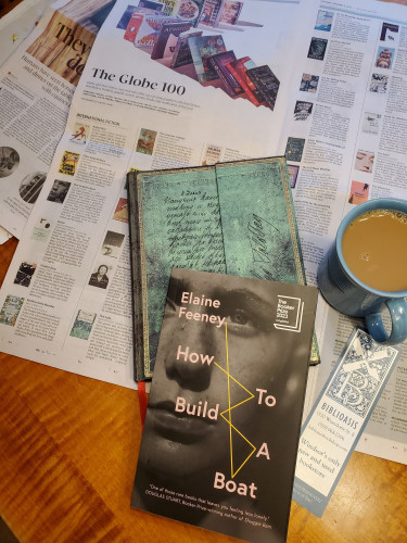 The novel How to Build a Boat by Elaine Feeney (Biblioasis) sits on a wooden table with a notebook with an ornate green cover, a Biblioasis bookmark, coffee in a blue cup and the Saturday Globe and Mail, showing their top 100 books of 2023, of which the Feeney book is one