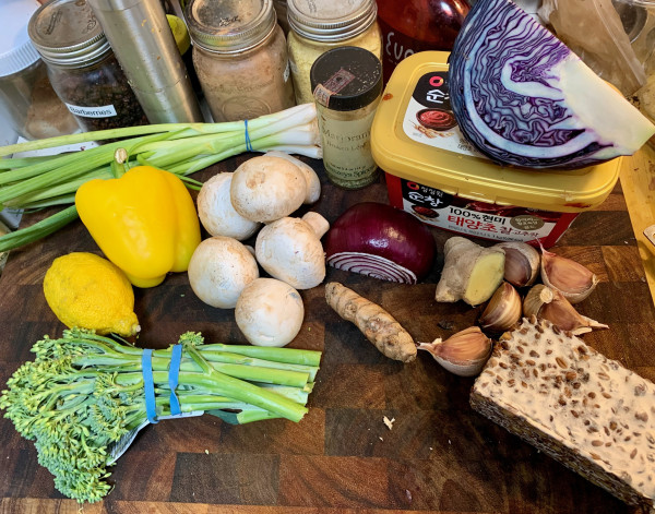 On a cutting board: 1 bunch of scallions, jar of dried marjoram, 1/4 red cabbage sitting on top of a tub of gochujanag, 5 large red garlicc cloves, block of tempeh, large turmeric root, small section of ginger root, 1/2 larg red onion, 6 medium white mushrooms, a bunch of broccolini, 1 lemon, 1 yellow bell pepper.