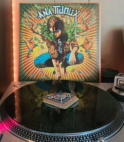 A black vinyl record sits on a turntable. Behind the turntable, a vinyl album outer sleeve is displayed. The front cover features art of Ana Tijou crouched down with tree branches coming out of her. There is a bird on each side of her. 