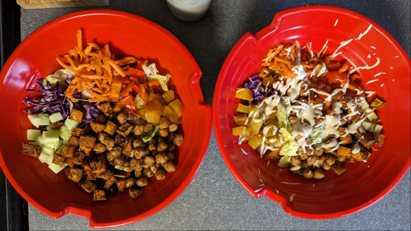 Two red bowls, the one on the right has a white dressing on the top. Bowls are filled with romaine lettuce, cucumber, red pepper,shredded purple cabbage,gold beet, roasted sweet potato, roasted chickpeas
