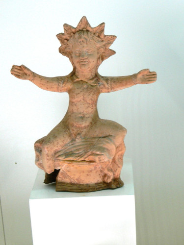 Small terracotta statuette of Helios with outstretched arms. He wears a sun ray crown and his arms are adorned with arm rings.