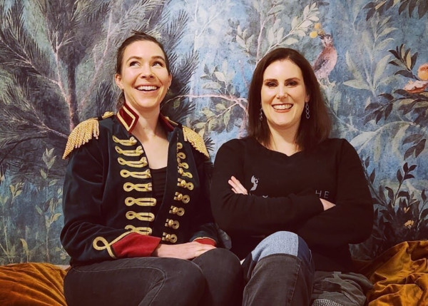 Dr G and Dr Rad sitting on a gold velvet lounge in front of a printed fresco from Livia’s garden villa. Both are smiling!