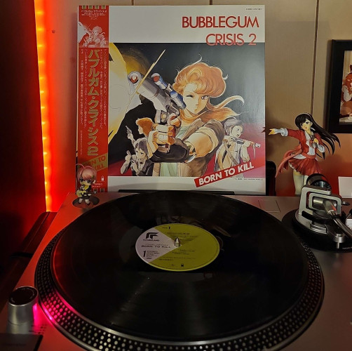 A black vinyl record sits on a turntable. Behind the turntable, a vinyl album outer sleeve is displayed. The front cover shows the 4 main cast members (Sylia, Priss, Linne, Nene) holding various guns. 