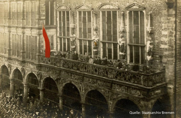 Eine historische Aufnahme des Bremer Rathauses. Auf dem Rathausbalkon stehen mehrere Leute, sie haben eine rote Flagge gehisst. Vor dem Rathaus auf dem Marktplatz steht eine dichtgedrängte Menschenmenge.