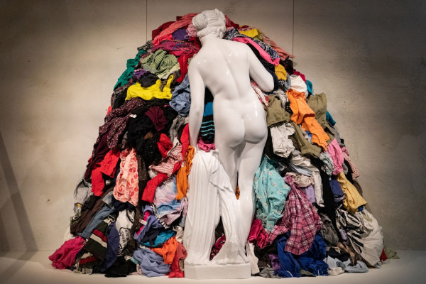 A photo showing a marble sculpture of a woman looking at a pile of clothes from throwaway fast fashion. This is part of the Throwaway exhibition at the House of European History, which explores the issue of waste in Europe from a historical perspective.