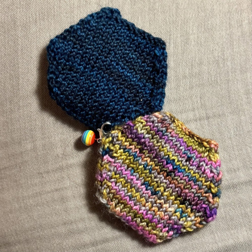 Hexiflats - knitted hexagon shapes - with two different yarns - tonal blue and multi-color. Blue hexiflat has a stitch marker with a rainbow striped ball attached.
