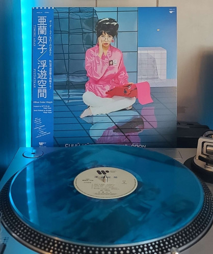 Image shows a turntable with a Transparent Blue vinyl record on the platter. Behind the turntable vinyl album outer sleeve is displayed. The front cover shows Tomoko Aran sitting cross legged on a floor with a typewriter on her lap. 