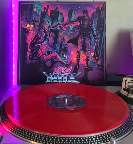 A Bloodshed Red vinyl record sits on a turntable. Behind the turntable, a vinyl album outer sleeve is displayed. The front cover shows a city, and in the street is a man in a leather coat pointing a gun down at a man begging for his life. 