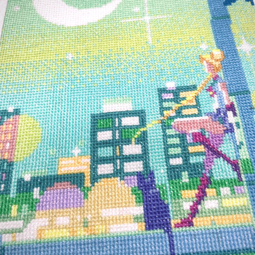 A cross stitch showing Sailor Moon and her cat Luna, watching the moon rising over the city night sky. The colors are a mix of greens and blues, with warm accent in pink and orange