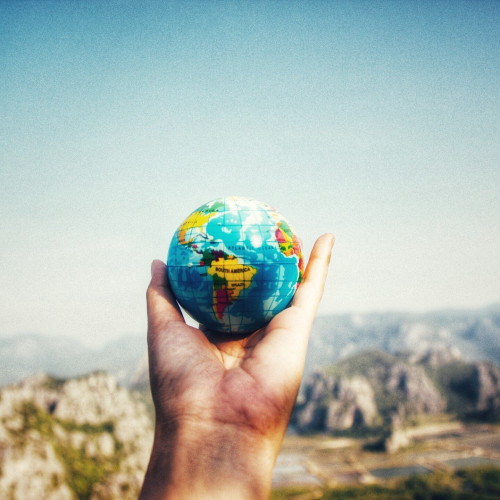 A person's hand holding a a small globe, with a rocky landscape and a blue sky horizon in the background.