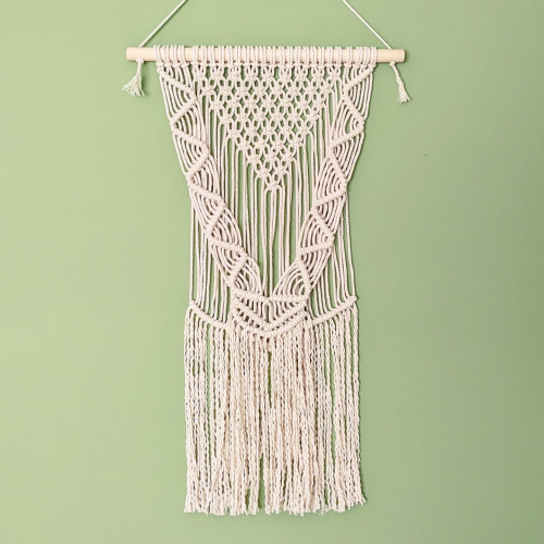 A completed macrame wall hanging in natural coloured cord, hanging against a green wall. The hanging consists of square knots in a triangular central arrangement, with densely-knotted V-shapes down each side meeting in the middle. The bottom half of the hanging is simple untwisted cord left to dangle freely.