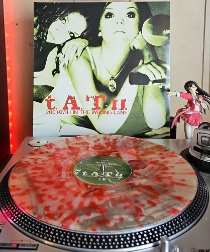 A clear w/ Red Splatter vinyl record sits on a turntable. Behind the turntable, a vinyl album outer sleeve is displayed. The front cover shows  the girls over tATu posing together on a motorcycle. 

To the right of the album cover is an anime figure of Yuki Morikawa singing in to a microphone and holding her arm out. 