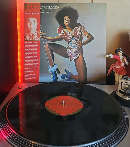 A black vinyl record sits on a turntable. Behind the turntable, a vinyl album outer sleeve is displayed. The front cover shows Betty Davis kneeling on one knee wearing an outfit that seems to be Egyptian inspired, and holding clear poles.. 

To the right of the album cover is an anime figure of Yuki Morikawa singing in to a microphone and holding her arm out. 
