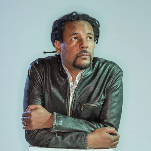 Portrait of author Colson Whitehead, a Black man with dreads tied in a ponytail. He wears a cool leather jacket. He gazes off to the upper right.