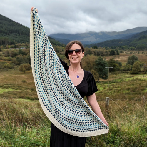 Me holding my finished handknit shawl, against a background of an autumnal Scottish glen. The shawl is half-circle shaped. It is white with rows of triangles in three different shades: light blue, mid-blue with silver, and dark blue. There is about 2" of white ribbing along the bottom.