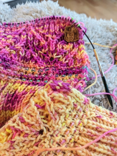 A brioche and slip stitch handknit shawl in progress in bright neon pinks and yellows with a few other colors hidden in the slip stitches. It has a cute Yarnicorn stitch marker on it.