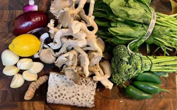 A wooden cutting board on which are seen a block of tempeh, a large turmeric root, six large garlic cloves, a lemon, 1/2 red onion, a pile of oyster mushrooms, three  jalapeños, a bunch of broccolini, and a large bunch of spinach.