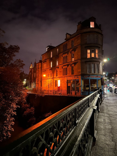 A view from a bridge across a river in a city. Along the other bank, a brick building, with a café on the ground floor and residences above, extends along the intersection between the road and the bank. The bankside wall of the building is illuminated by a bright orange street lamp, whose light reflects from a railing nearer the camera. Lights along the road are a whiter shade. It has rained recently and the wet ground reflects the light.