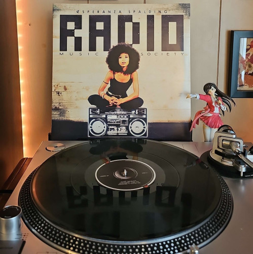 A black vinyl record sits on a turntable. Behind the turntable, a vinyl album outer sleeve is displayed. The front cover shows Esperanza Spalding sitting on a boombox. 

To the right of the album cover is an anime figure of Yuki Morikawa singing in to a microphone and holding her arm out. 
