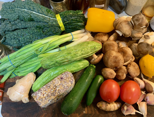 A cutting board with a large ginger root, 2 bunches thick scallions, a bunch of black kale, yellow bell pepper, oyster mushrooms, shiitake mushrooms, bitter melon tempeh, green zucchini, green jalapeño, thick turmeric root, 2 tomatoes, 6 cloves red garlic, and a lemon. It's a crowded cutting board.