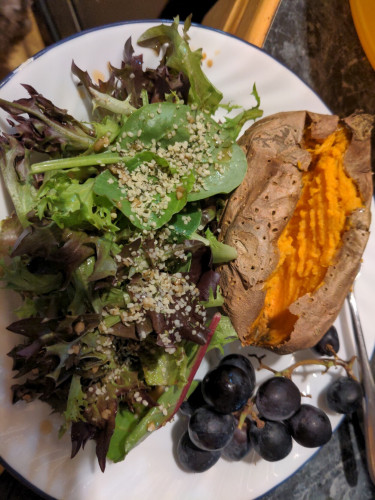 A plate of black grapes, baked sweet potato, and greens with dressing and hemp seeds.