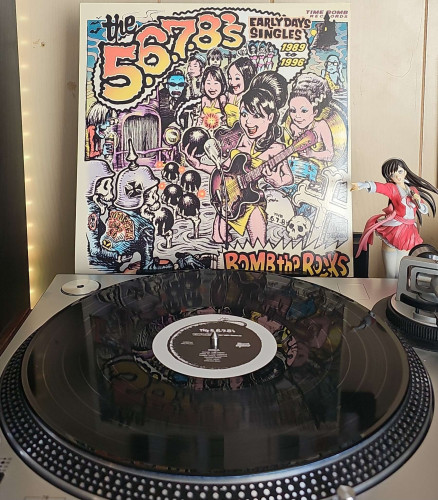 A black vinyl record sits on a turntable. Behind the turntable, a vinyl album outer sleeve is displayed. The front cover shows comic book style characters of The 5, 6,7 , 8s and various monsters. 

To the right of the album cover is an anime figure of Yuki Morikawa singing in to a microphone and holding her arm out. 