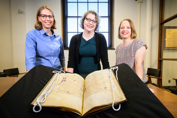 Three people standing behind a large open manuscript.