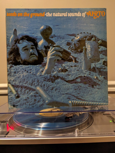 Seeds on the Ground: The Natural Sounds of Airto LP. Album cover w/ photo of Airto buried in the sand with percussion instruments. Ocean blue vinyl.