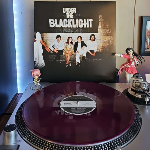 A Purple Translucent vinyl record sits on a turntable. Behind the turntable, a vinyl album outer sleeve is displayed. The front cover shows Rilo Kiley sitting in front of a brick wall that has Under The Blacklight painted on it. 