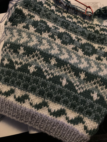 The back piece of a baby jumper knitted in multiple bands of Fair Isle knitting, which use three shades of wool (sage, darker more teal green and white) - the ribbed cuffs will be in a bluey lilac colour 