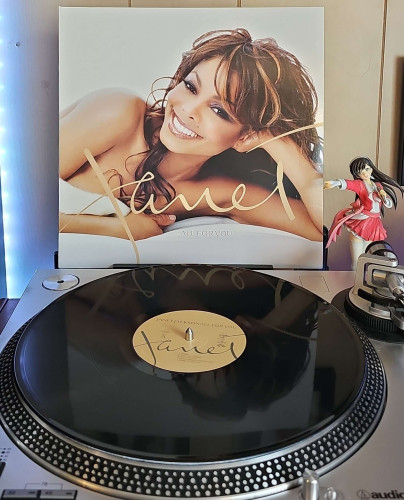 A black vinyl record sits on a turntable. Behind the turntable, a vinyl album outer sleeve is displayed. The front cover shows Janet Jackson laying down and smiling at the camera. 

To the right of the album cover is an anime figure of Yuki Morikawa singing in to a microphone and holding her arm out. 
