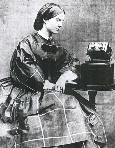 A black and white photograph of Mary Ward. She wears a dress and is sitting at a small table. At the table, there is want appears to be some sort of microscope (I think? I'm not sure). She looks down at the device in a contemplative manner.