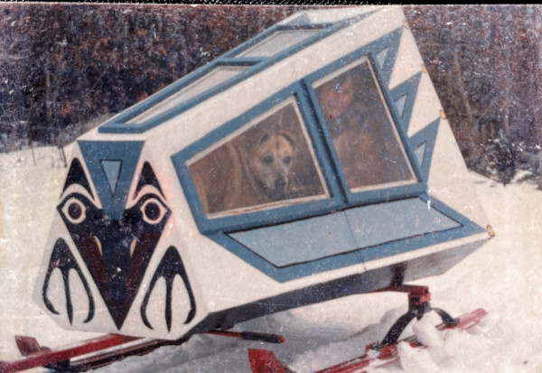 A closed plywood snowmobile trailer (kind of like a rectangular funnel shape) painted with a stylistic Thunderbird. Through the window you can see my mother and her dog.