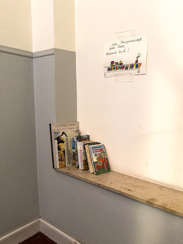 A group of book on a stone board in front of a gray and white painted wall within a staircase. A handwritten sign with painted books in a shelf. The sign reads:
de:
"Liebe Hausgemeinschaft und Gäste. 
Bedient euch!"
en:
"Dear house community and guests. 
Help yourselves!"