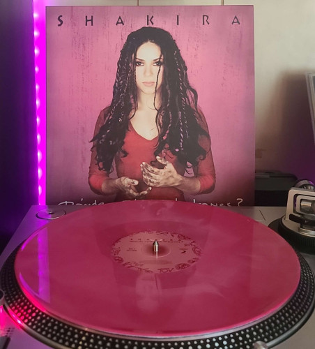 A Red & Pink Galaxy vinyl record sits on a turntable. Behind the turntable, a vinyl album outer sleeve is displayed. The front cover shows Shakira looking at the camera. She is holding her dirty hands in front of her just away from her shirt.