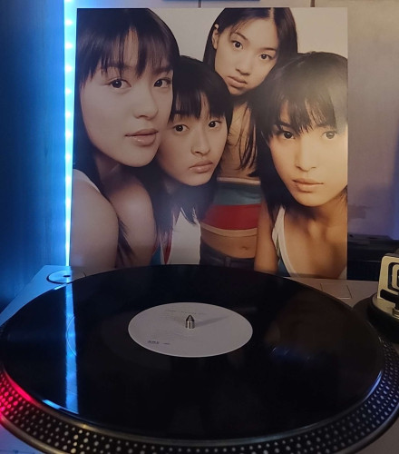 A black vinyl record sits on a turntable. Behind the turntable, a vinyl album outer sleeve is displayed. The front cover shows the 4 members of Speed huddled together looking at the camera. 