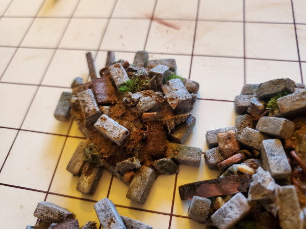 A picture of three pieces of terrain for DnD. They're rubble piles, between 2-6 inches across. They're made of little bricks, wooden boards, chains, moss, some, grass bits, and other detritus.