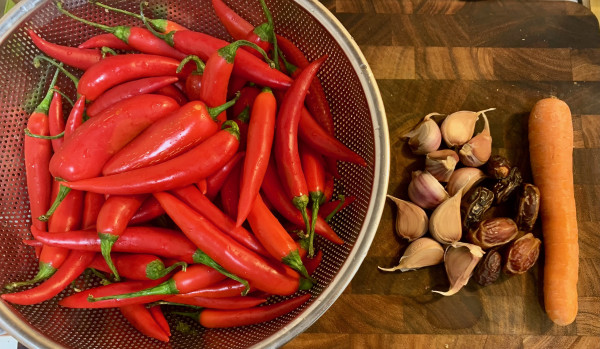 A collander holding cayenne peppers and a few red jalapeño peppers on a cutting board next to 10 large cloves red garlic, 7 Medjool dates, and a carrot.