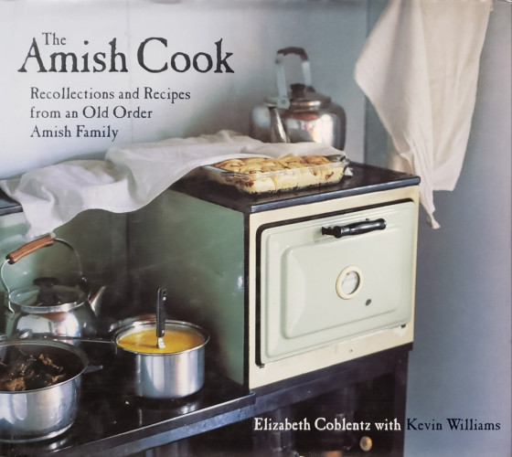 A photo of a book.

The Amish Cook – Recollections and Recipes from an Old Order Amish Family.
Elizabeth Coblentz with Kevin Williams. 

The photo on the cover is a very nice but very old stove and oven. A pie with a cloth loosely draped over it rests, cooling, in a rectangular glass baking pan atop the oven. Behind it sits a large silvery kettle. To the left, onf back burner of the shorter surface for the stove holds another, smaller, silver kettle with a wood handle. There are two shiny metal pots up front. One appears to contain meat, the other contains something of a yellow-orange hue with a utensil handle sticking up and standing upright in the center.