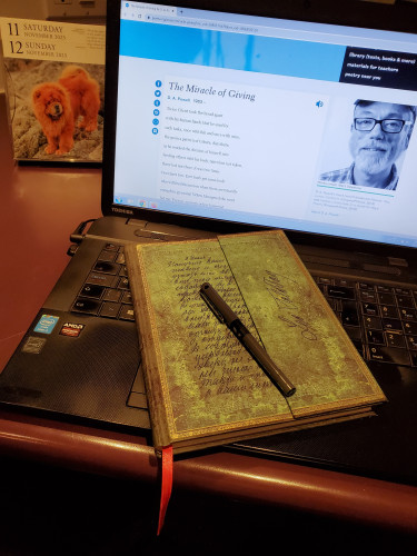The poem "The Miracle of Giving" by D.A. Powell appears on a computer screen on the web site of Academy of American Poets, with a notebook with an ornate green cover sitting on the computer keyboard. A dog calendar is in the background.