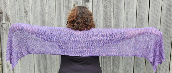 A woman with shoulder length brown curly hair is facing a barn while holding a long knit Shawl across her back. The Shawl is in a lovely purple variegated yarn and completely facing the camera. The pattern uses a palmette motif common to Egyptian, Assyrian, Phoenician, Cypriote and Greek Art.

