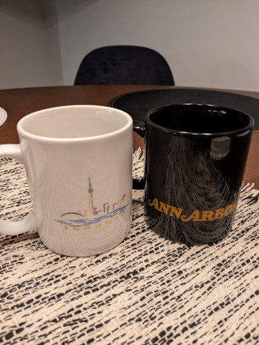 A white coffee mug with an abstract drawing of the skyline of Toronto on it (with the text Toronto below) and a black coffee mug that says Ann Arbor (a city in Michigan)